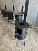 Fogão a lenha industrial preto com tampa e alças amarelas, em piso rústico, contra parede cinza