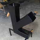 Churrasqueira de aço preta inclinada com cilindros de madeira, sobre piso cinza