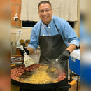 Homem com avental preto preparando comida em chapa grande com linguiça e arroz em ambiente interno